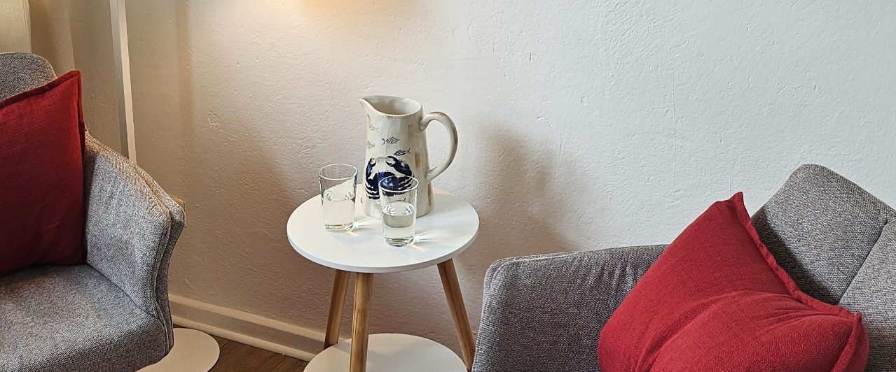 EmpathieundMeer(6) Blick in die Praxis auf die zwei Stühle mit weinroten Kissen, Beistelltisch mit Wasserkaraffe und zwei Gläsern