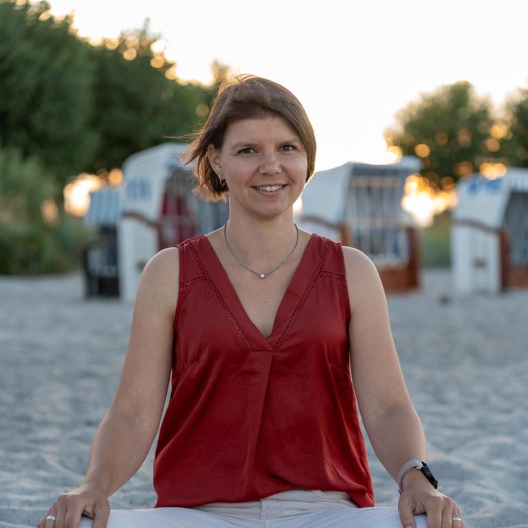 Sophie Rödiger sitzt lächelnd am Strand, im Hintergrund Strandkörbe und untergehende Sonne